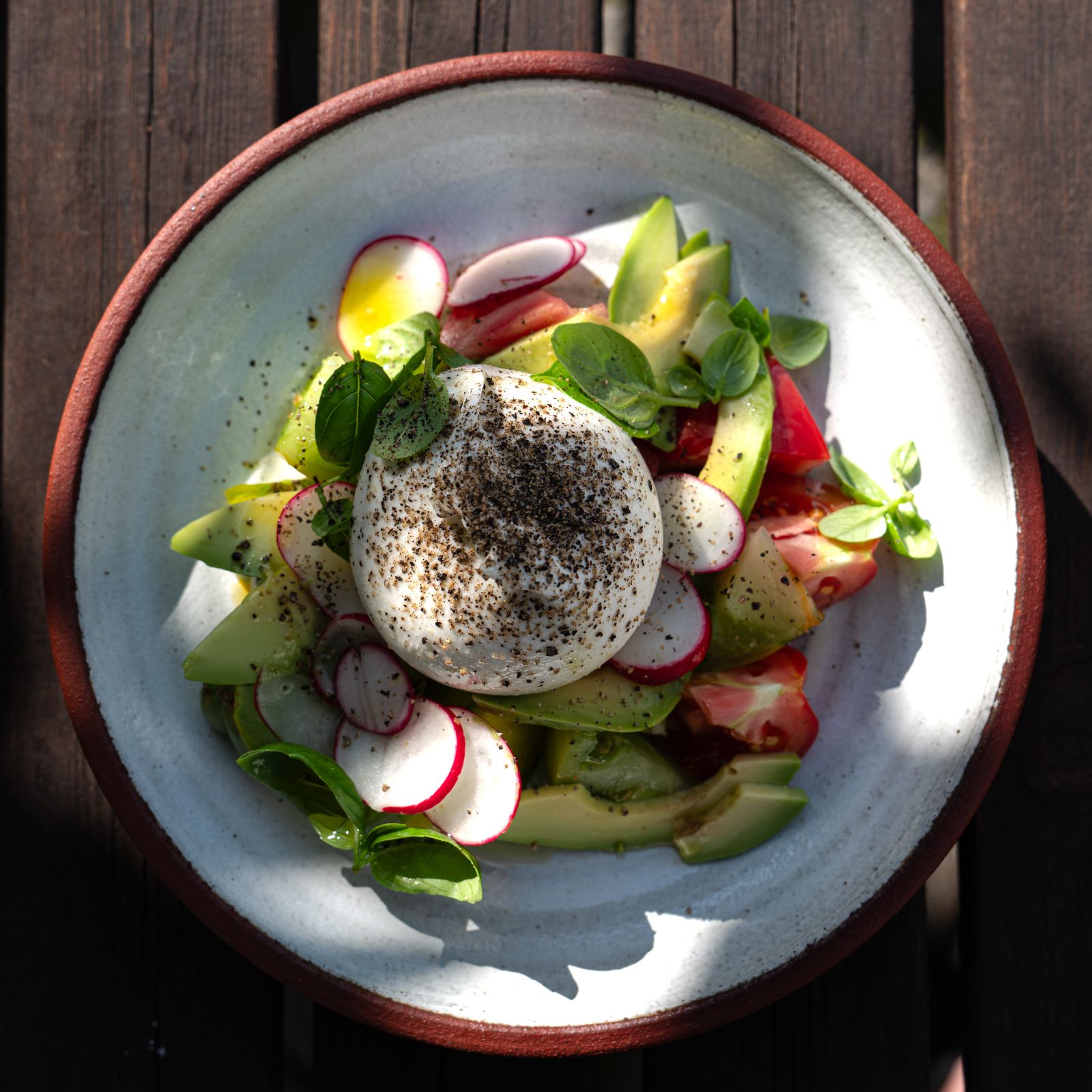Delicious burrata salad with fresh basil, tomatoes, radishes, and avocado on a rustic wooden table.